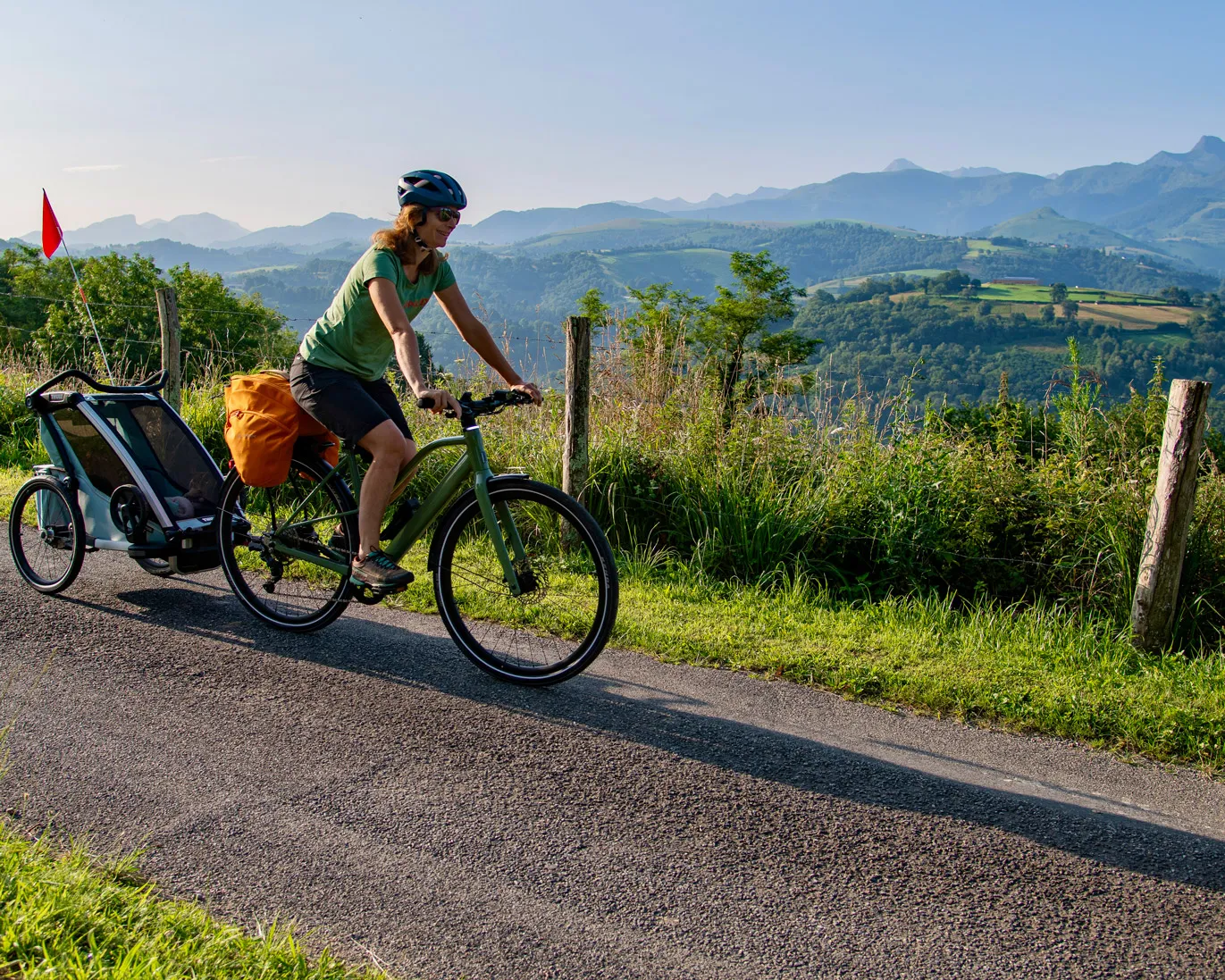 Femme à vélo portant un casque, tirant une remorque, sur une route de campagne avec montagnes en arrière-plan.