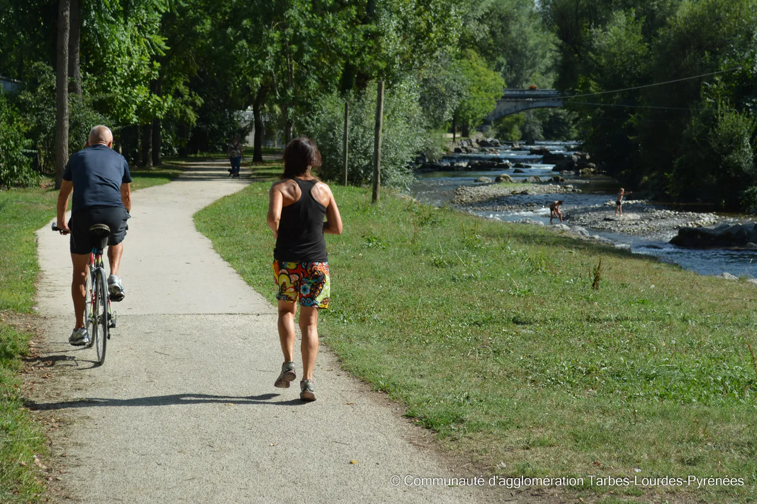 Un homme à vélo et une femme courant sur un chemin près d'une rivière avec des arbres et un pont.