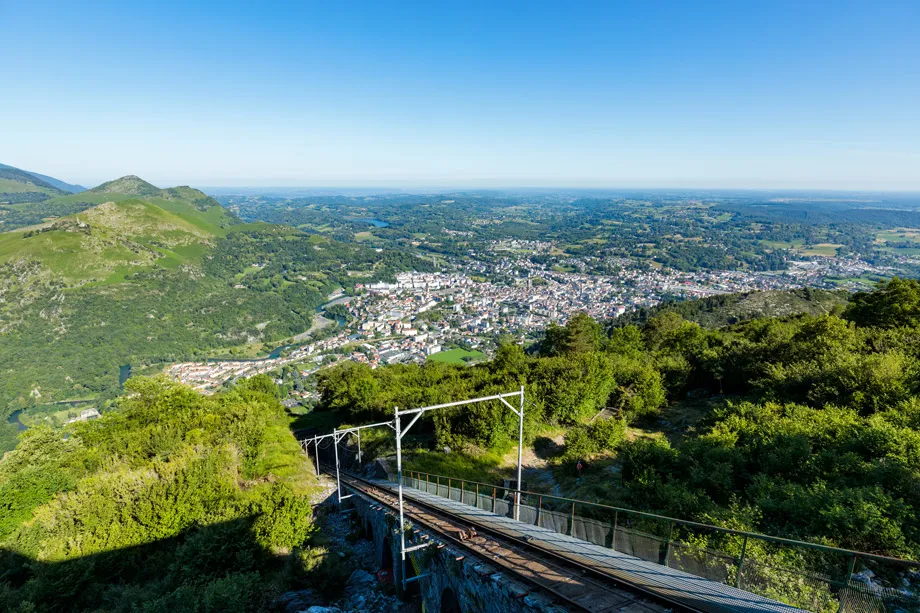 Vue panoramique d'un funiculaire descendant une montagne verte vers une ville en contrebas par temps clair.