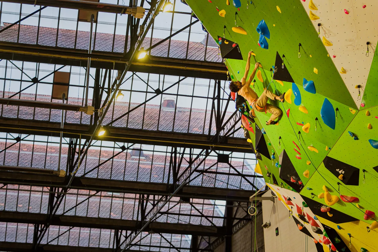 Un grimpeur sur un mur d'escalade intérieur avec prises colorées sous une verrière en acier.