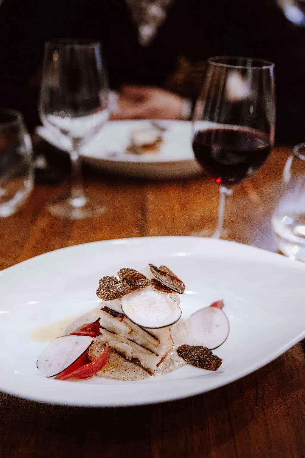 Plat gastronomique avec tranches de truffe et légumes sur une assiette blanche, verre de vin rouge en arrière-plan.