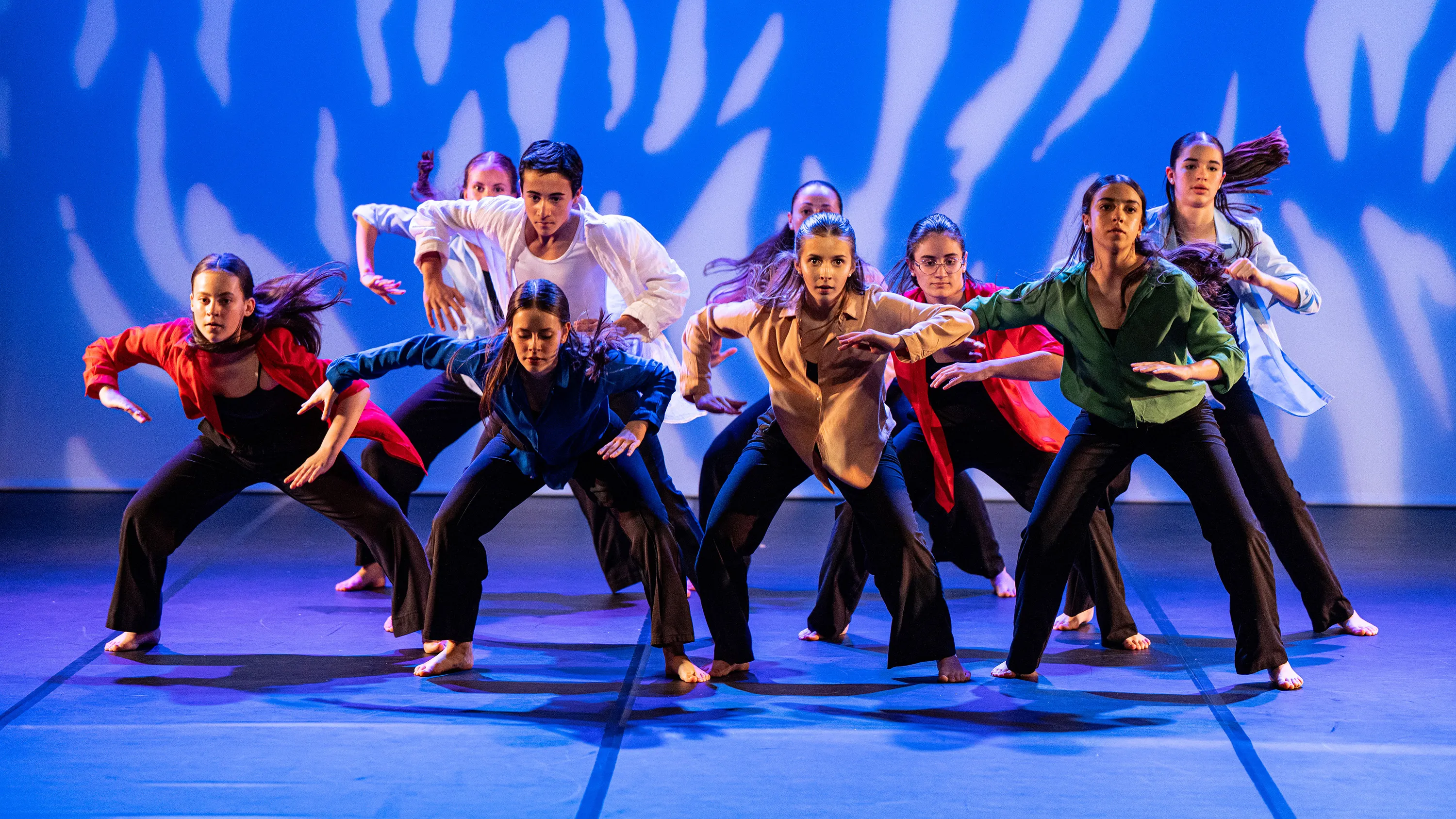 Groupe de danseurs en chemises colorées et pantalons noirs dans une pose synchronisée sur scène.