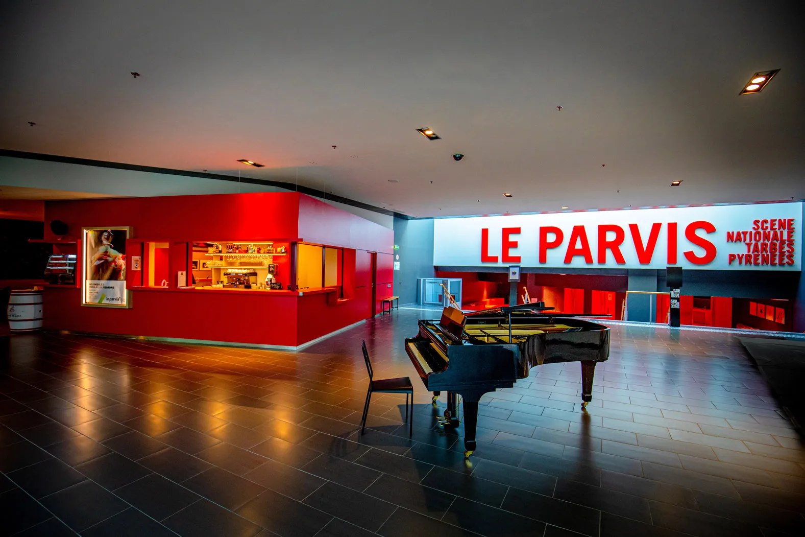 Hall intérieur avec un piano à queue noir, un comptoir rouge et un grand panneau lumineux « LE PARVIS ».