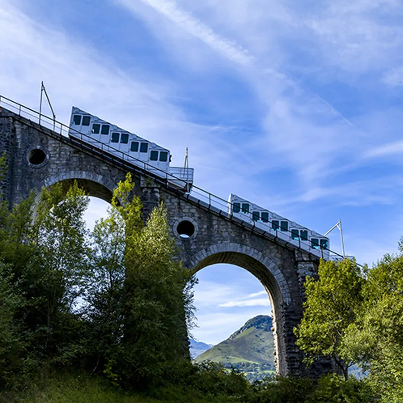 Un funiculaire transporte des passagers sur un pont-viaduc en pierre dans un paysage montagneux vert.