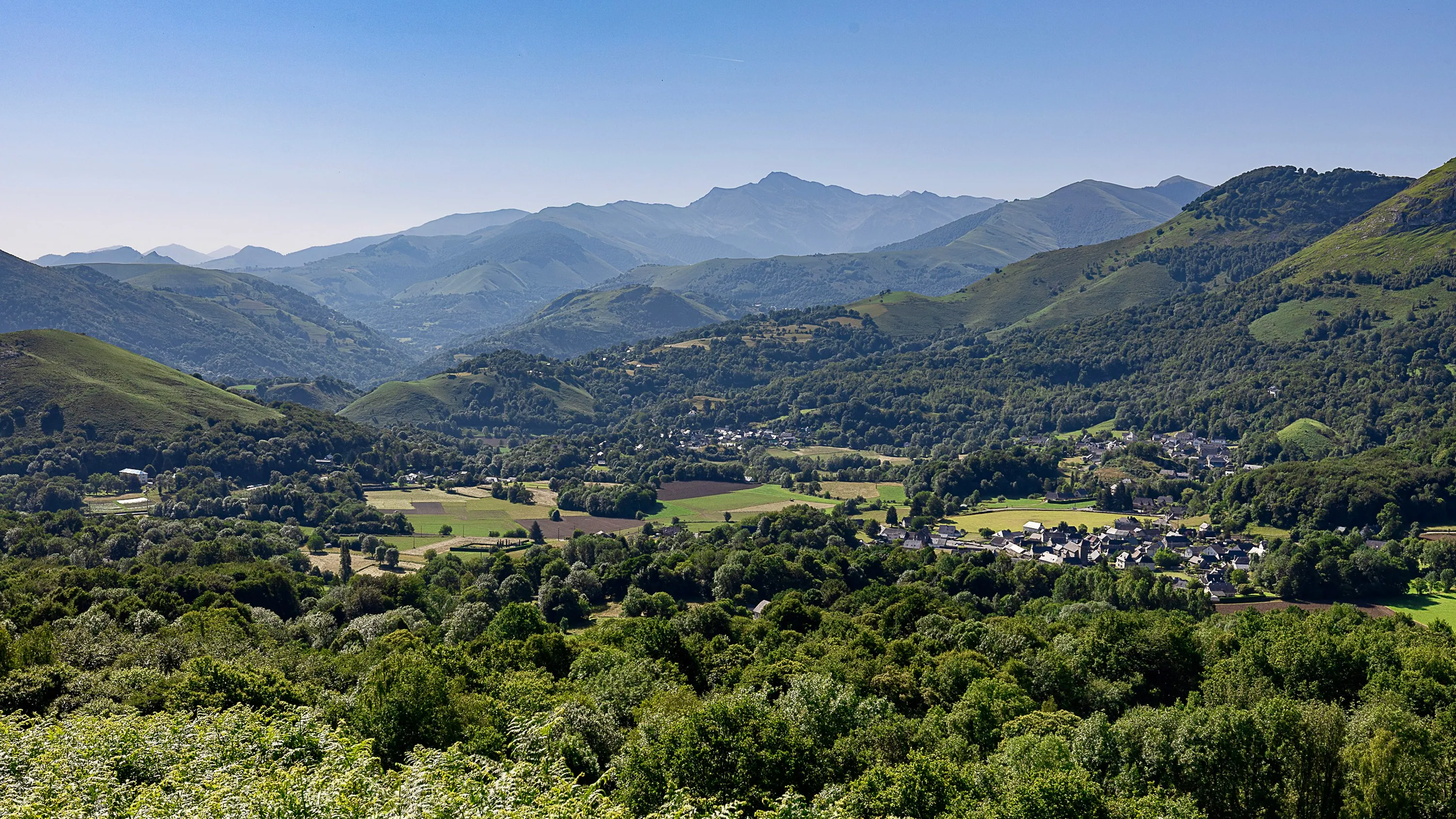 Vallée verdoyante entourée de collines et montagnes sous un ciel bleu clair.