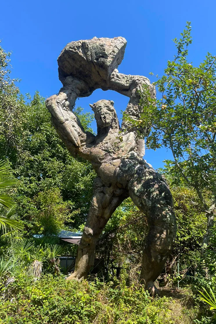 Statue imposante d'un géant en pierre tenant une grande roche au-dessus de sa tête dans un jardin.