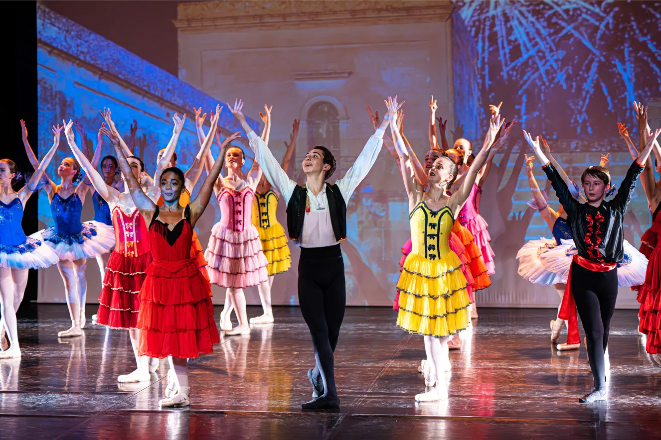 Groupe de jeunes danseurs de ballet en costumes colorés sur scène, bras levés en position finale.