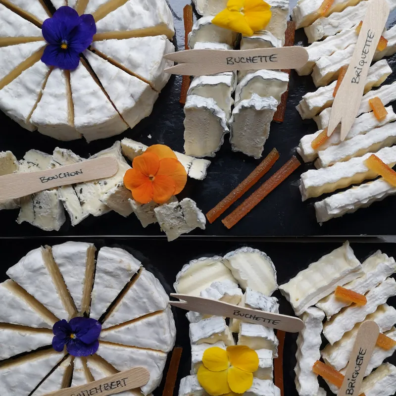 Plateau de fromages variés tranchés avec fleurs comestibles et étiquettes en bois.
