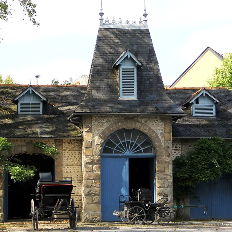 Bâtiment en pierres avec porte bleue, toits en ardoise et carrioles anciennes devant l'entrée.