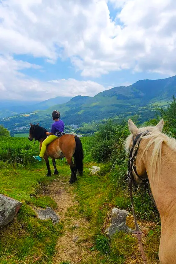 Enfant à cheval sur sentier de montagne, vue sur vallées et montagnes sous ciel nuageux.