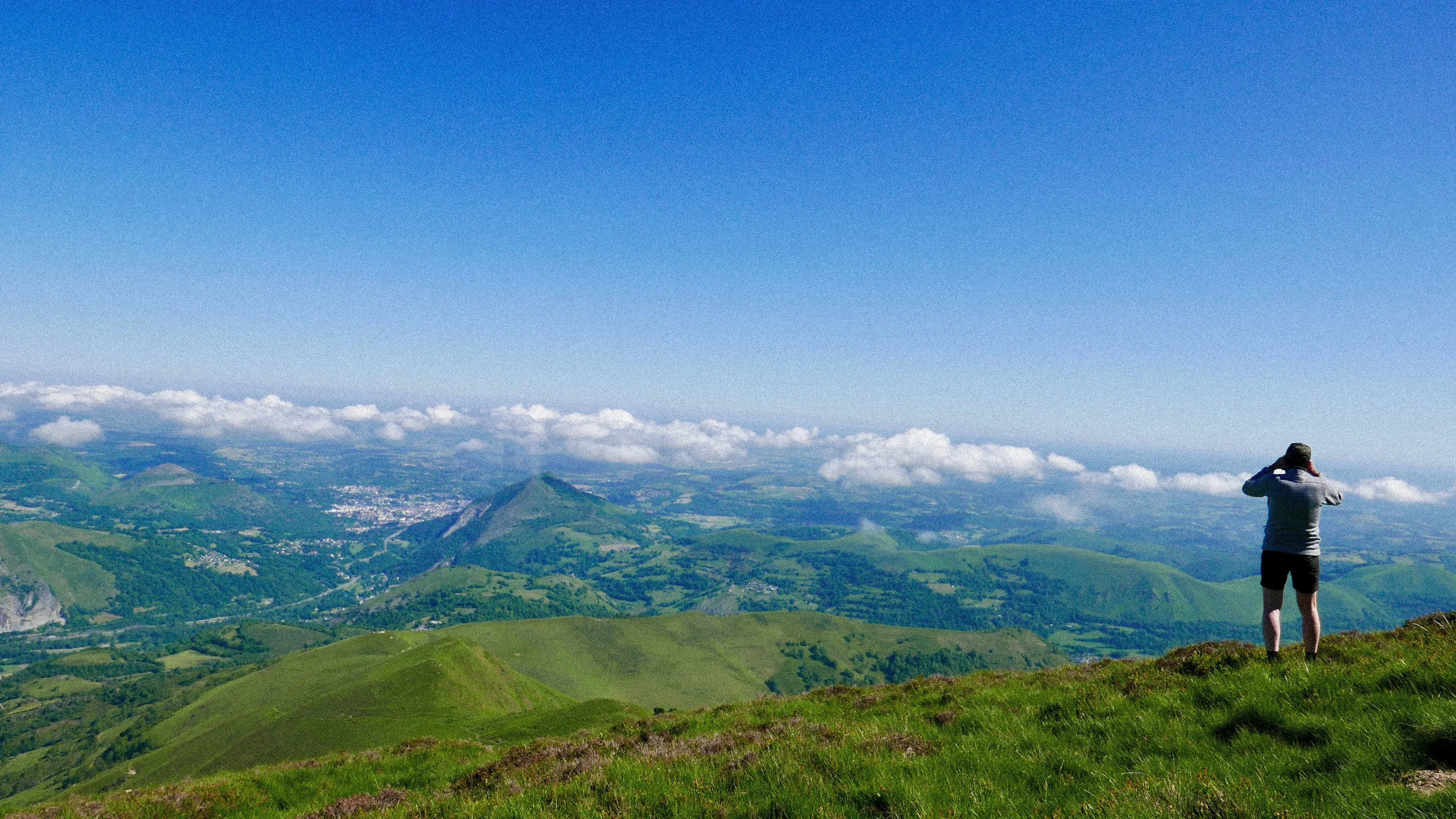 Une personne debout en short et pull gris regarde à travers des jumelles une vaste vallée verte avec des collines et un ciel bleu parsemé de nuages.