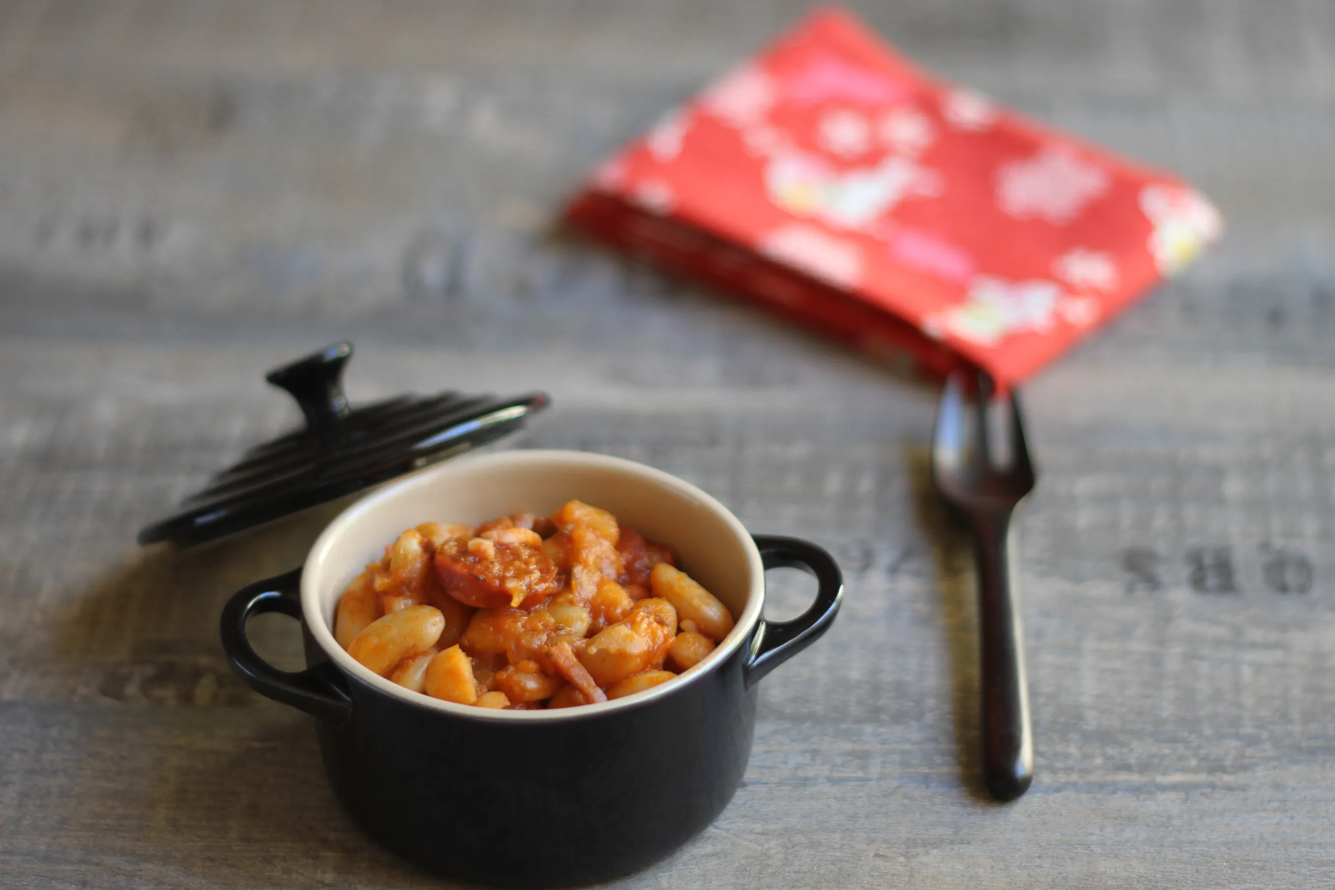 Petit cocotte noire remplie de haricots blancs au chorizo dans une sauce tomate, avec une fourchette.
