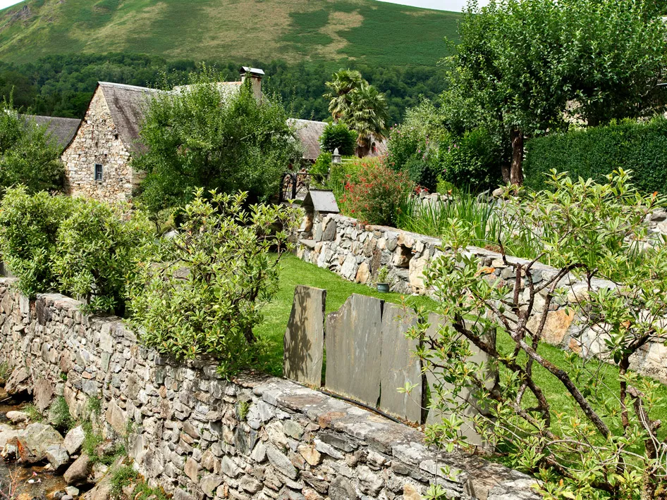 Jardin avec des murs en pierre, plantes, arbres, maisons en pierre et colline verte en arrière-plan.