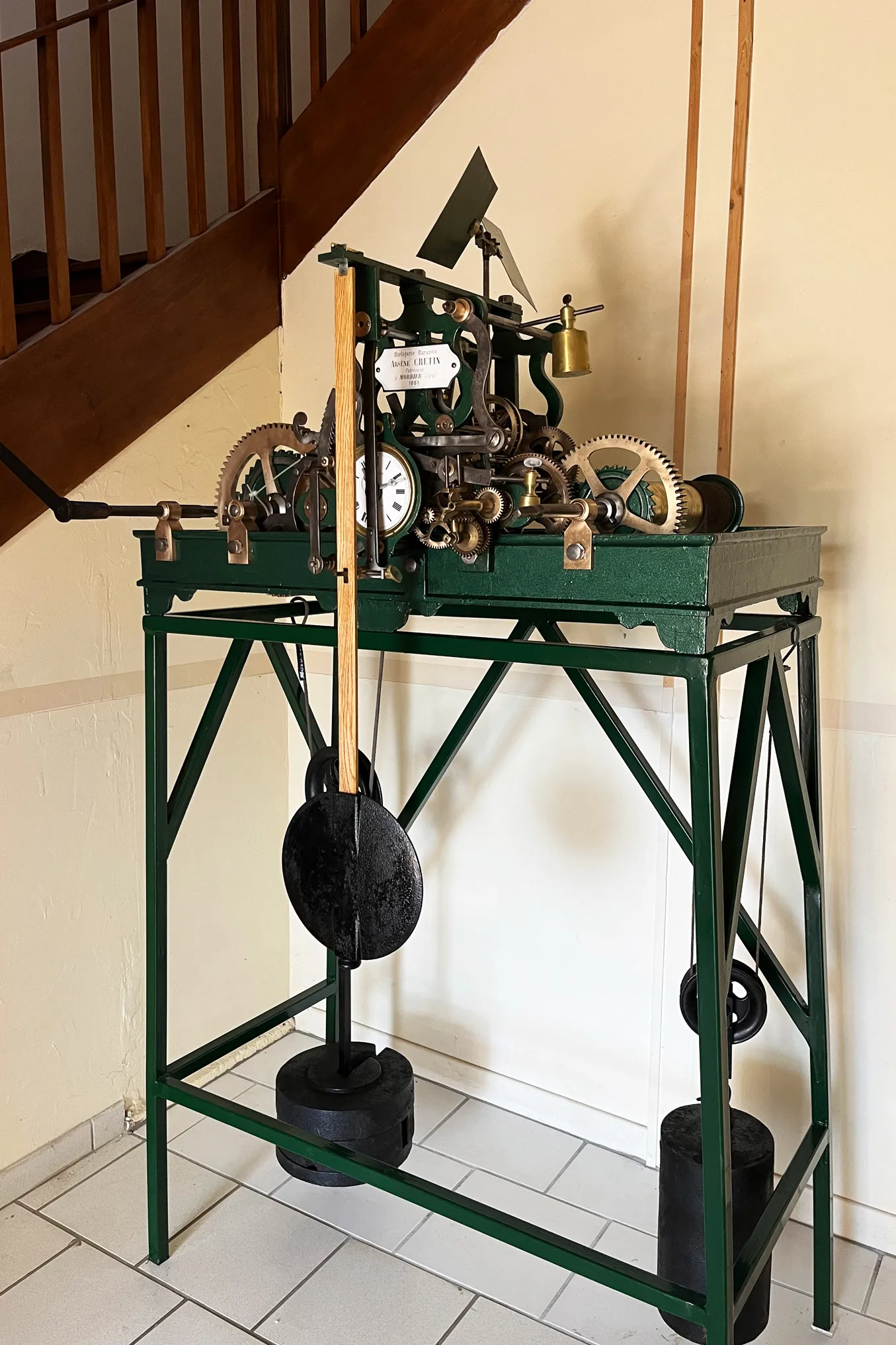 Ancienne horloge mécanique avec engrenages, poids et balancier sur un support métallique vert.