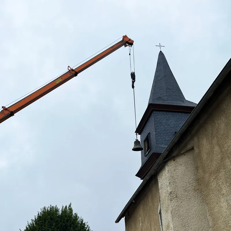 Une grue orange soulève une cloche près du clocher d'une église sur un fond de ciel gris.