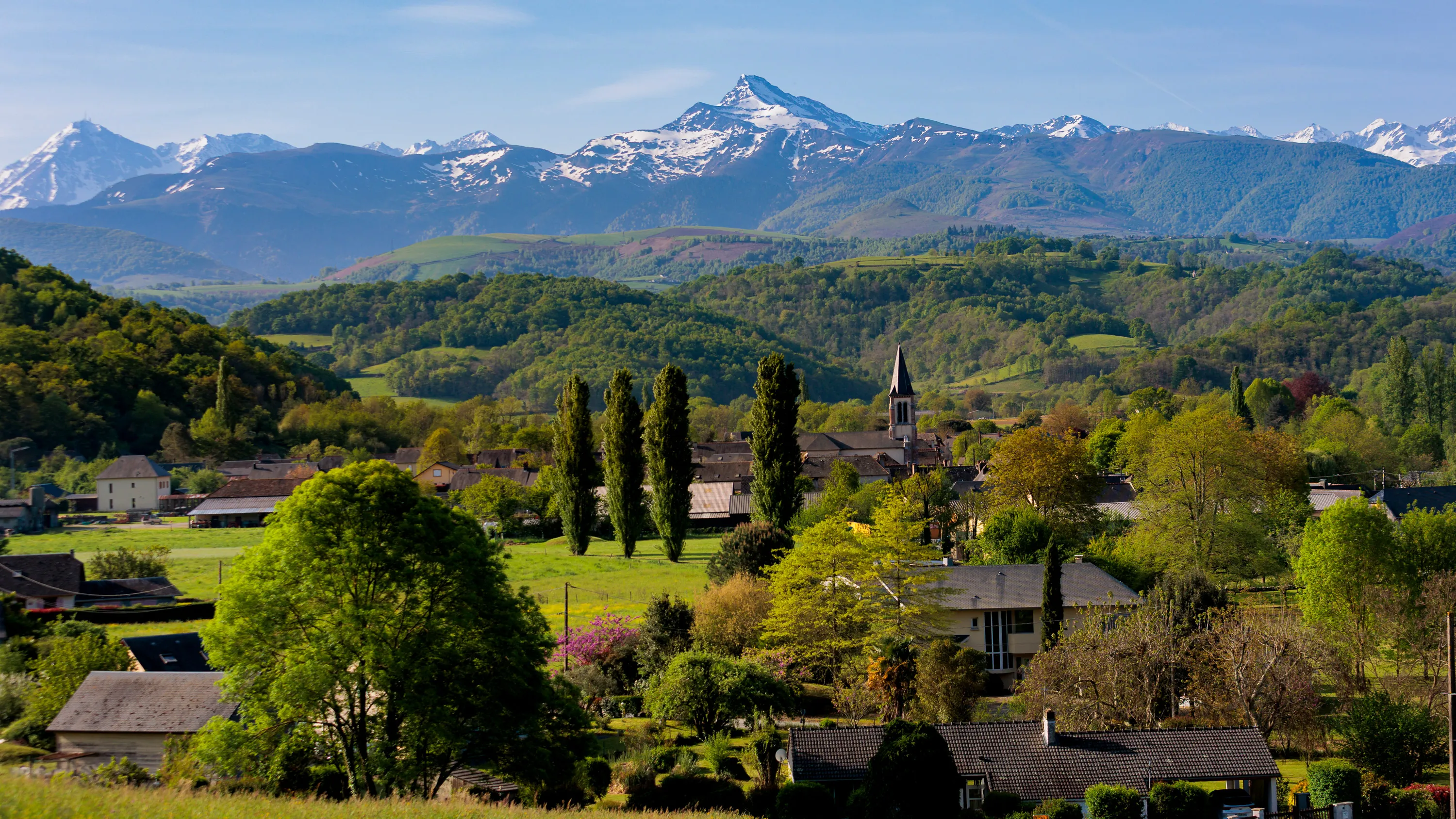 Village entouré d'arbres verts avec montagnes enneigées au loin sous un ciel bleu clair.