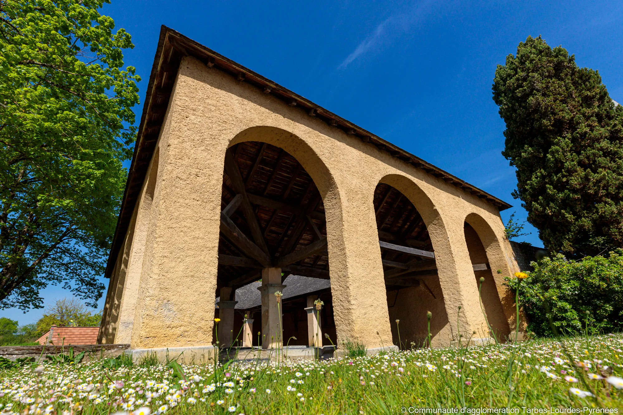 Lavoir en pierre avec arcs ouverts entouré d'herbe et de fleurs sous un ciel bleu clair.