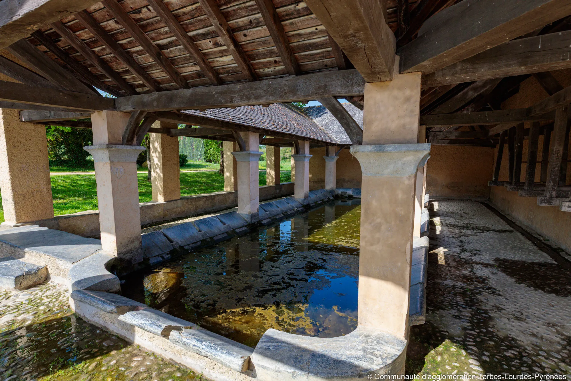 Ancien lavoir sous un toit en bois avec eau stagnante et piliers en pierre à la campagne.