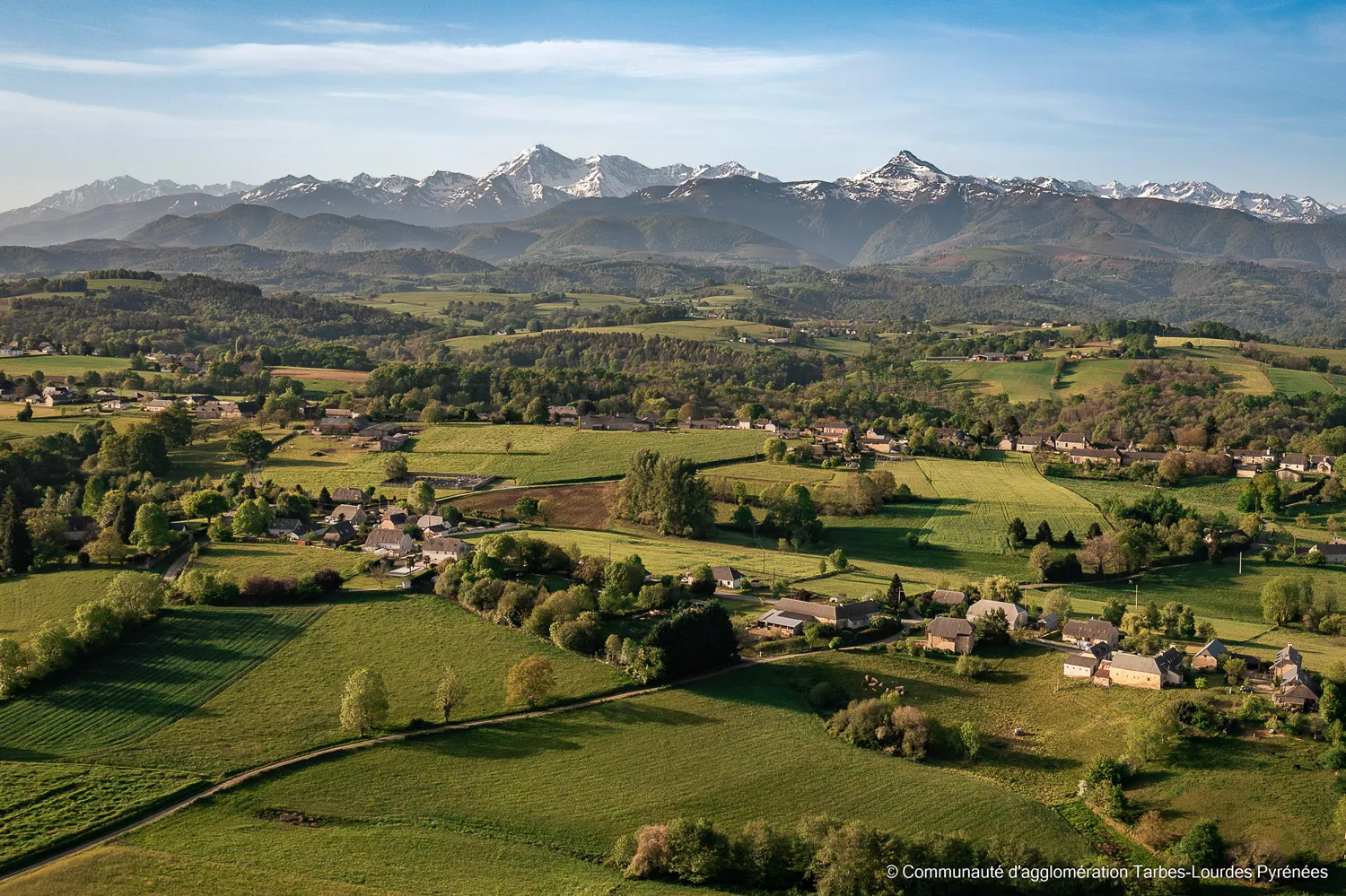 Vue aérienne d'un paysage rural avec des champs verts et des montagnes enneigées en arrière-plan.