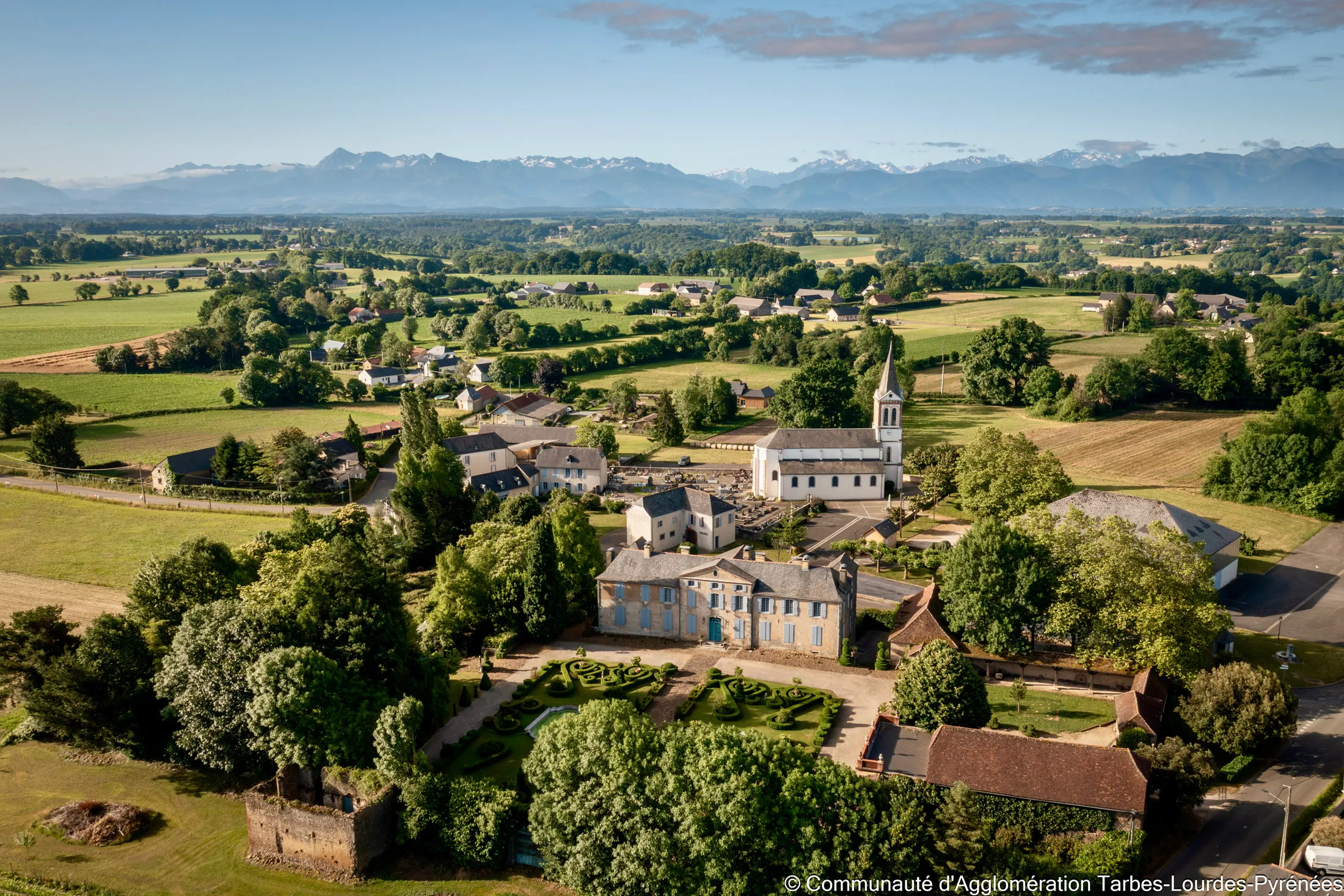 Vue aérienne de Garderes, un village avec un jardin à la française, une église et des montagnes en arrière-plan.