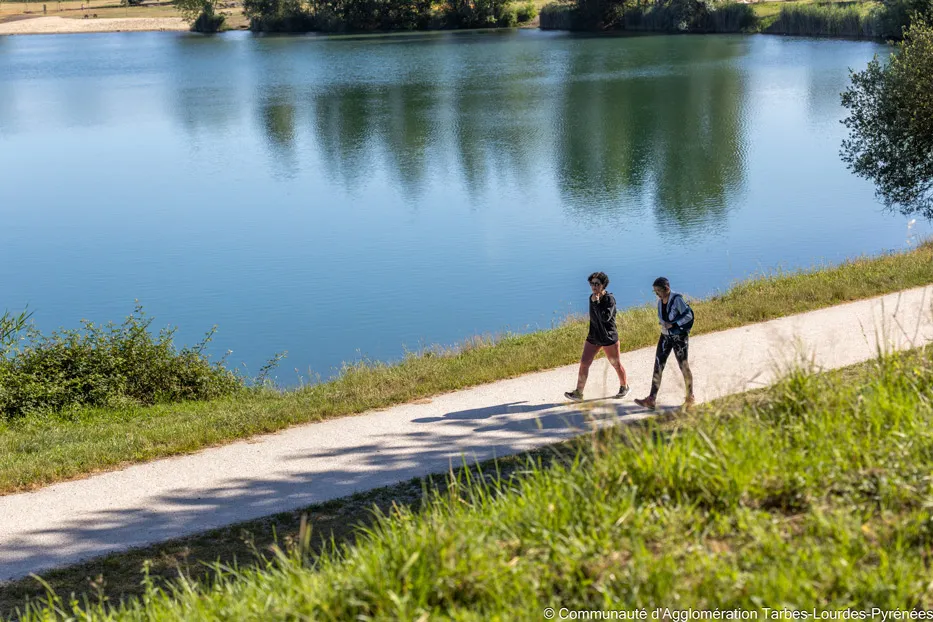 Deux personnes marchent le long d'un sentier au bord d'un lac calme avec des arbres en arrière-plan.