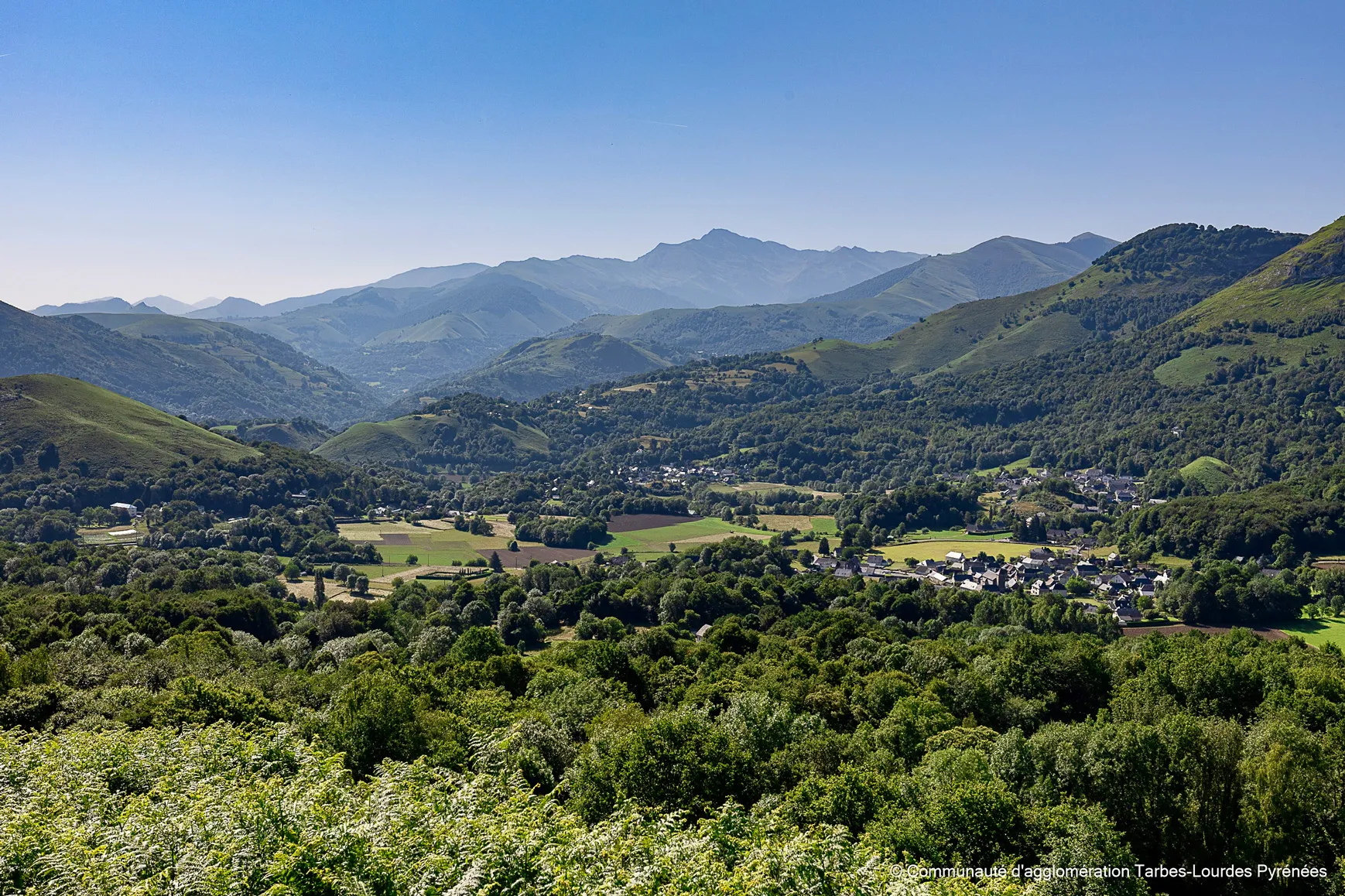 Vue panoramique de la vallée verte entourée de collines et montagnes sous un ciel bleu clair.