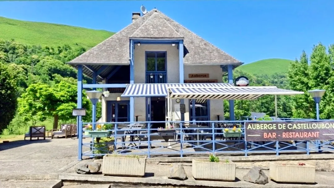 Façade de l'auberge de Castelloubon avec terrasse et store rayé bleu et blanc en milieu de verdure.
