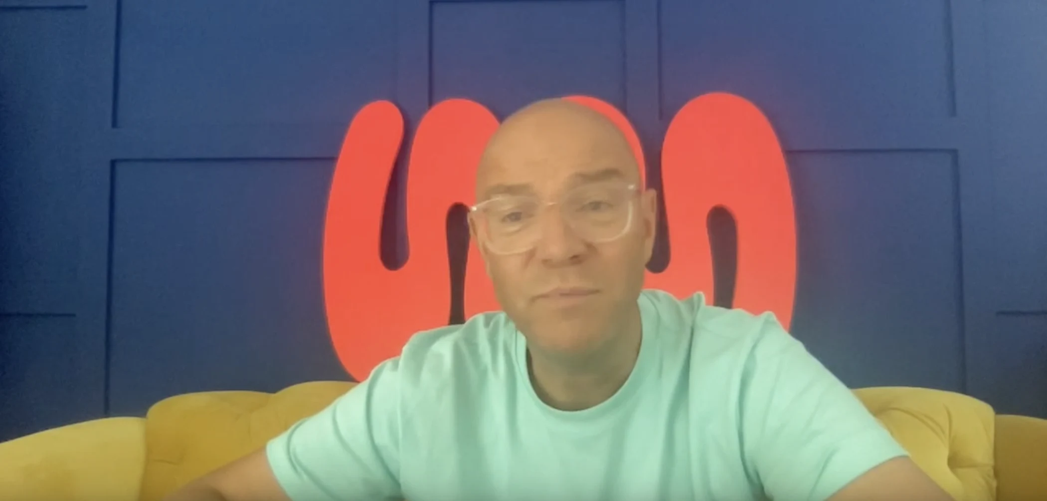 Bald man wearing glasses and a light blue shirt sitting on a yellow couch with a dark blue wall and red abstract shapes behind him.