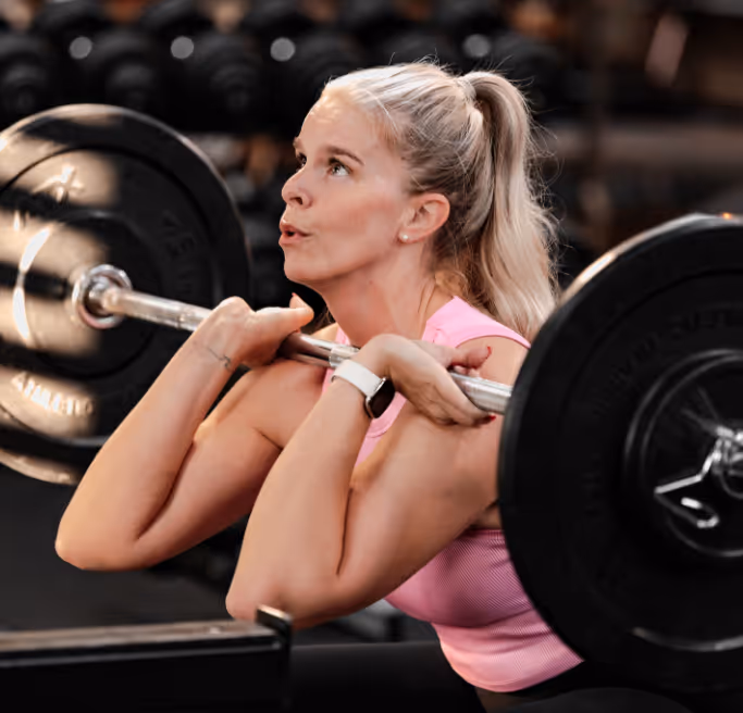 Woman in pink tank top lifting a barbell at a gym with focused expression.