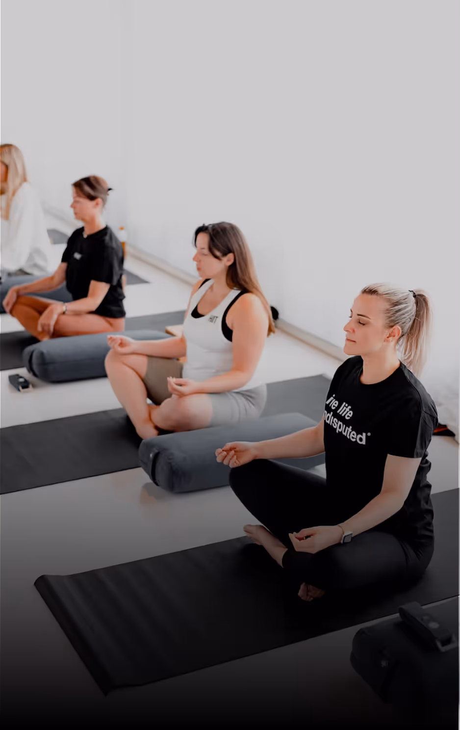 Four women sitting cross-legged on yoga mats in a meditation pose with eyes closed in a bright room.