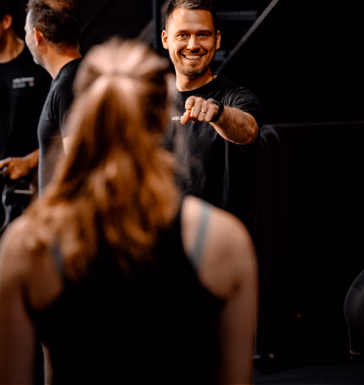 A smiling man points forward while engaging with a woman whose back is to the camera in a dimly lit setting.