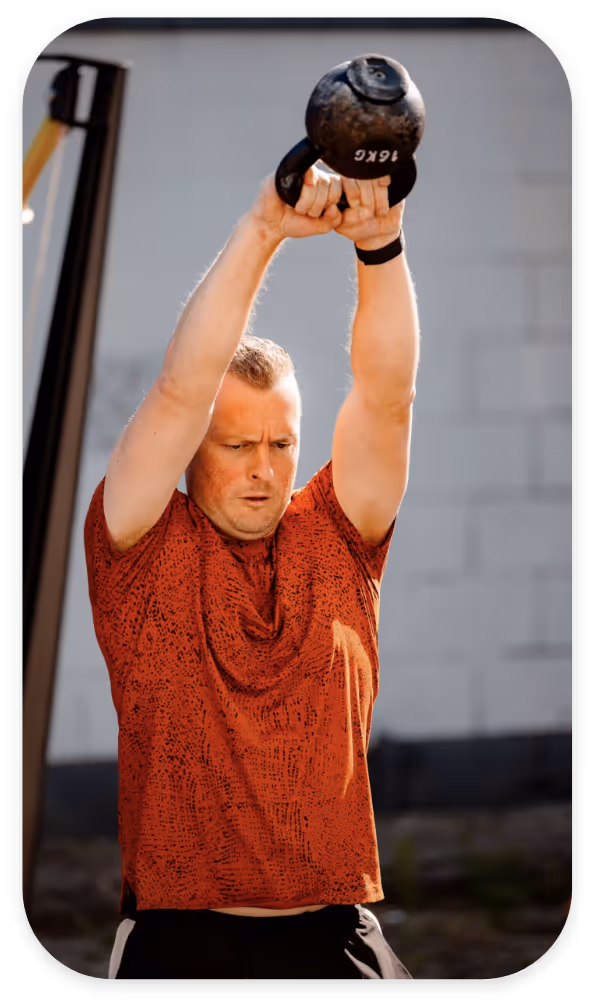 Man in red shirt lifting a 16kg kettlebell overhead during outdoor workout.