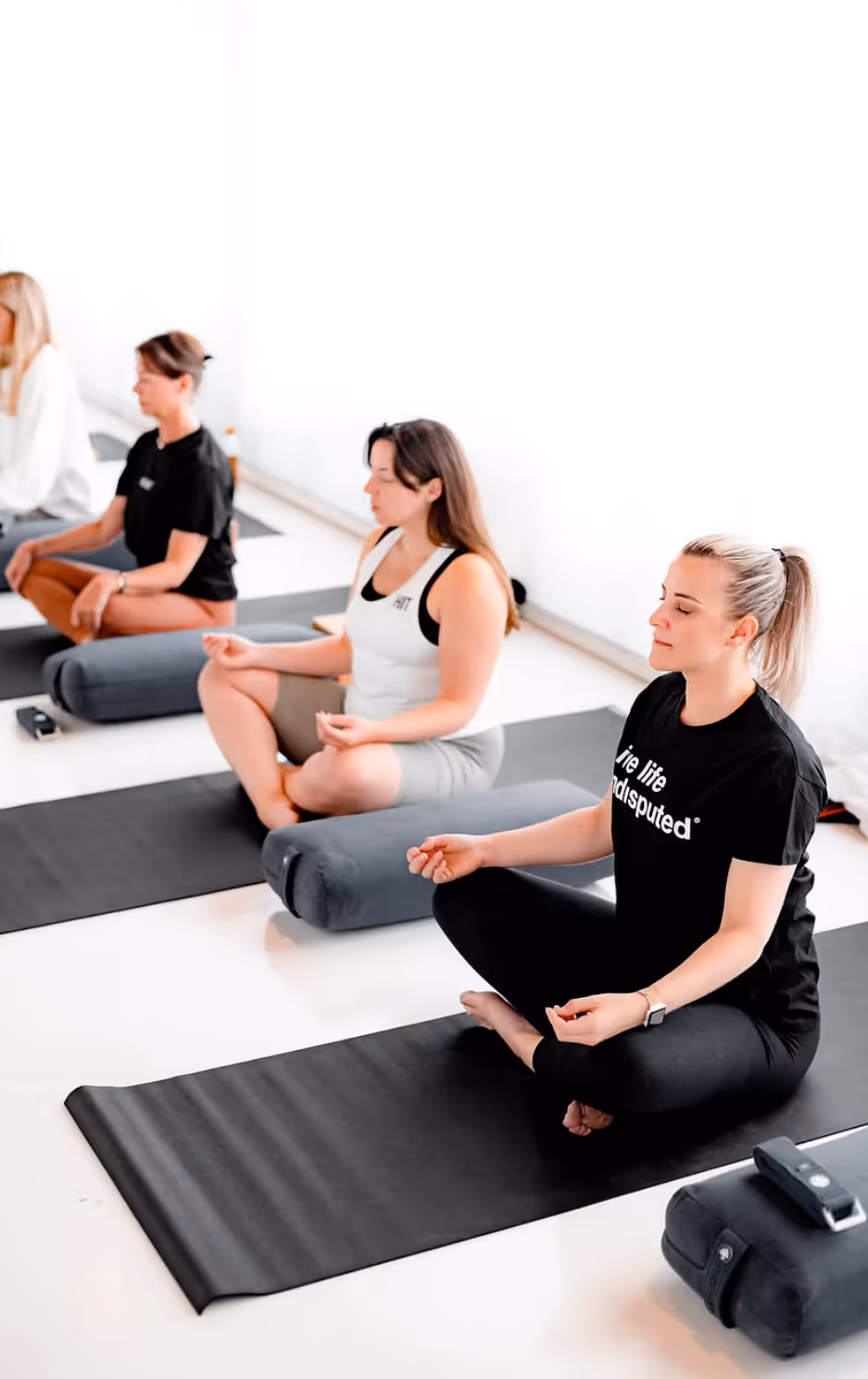 Four women sitting cross-legged on yoga mats in a meditation pose with eyes closed.