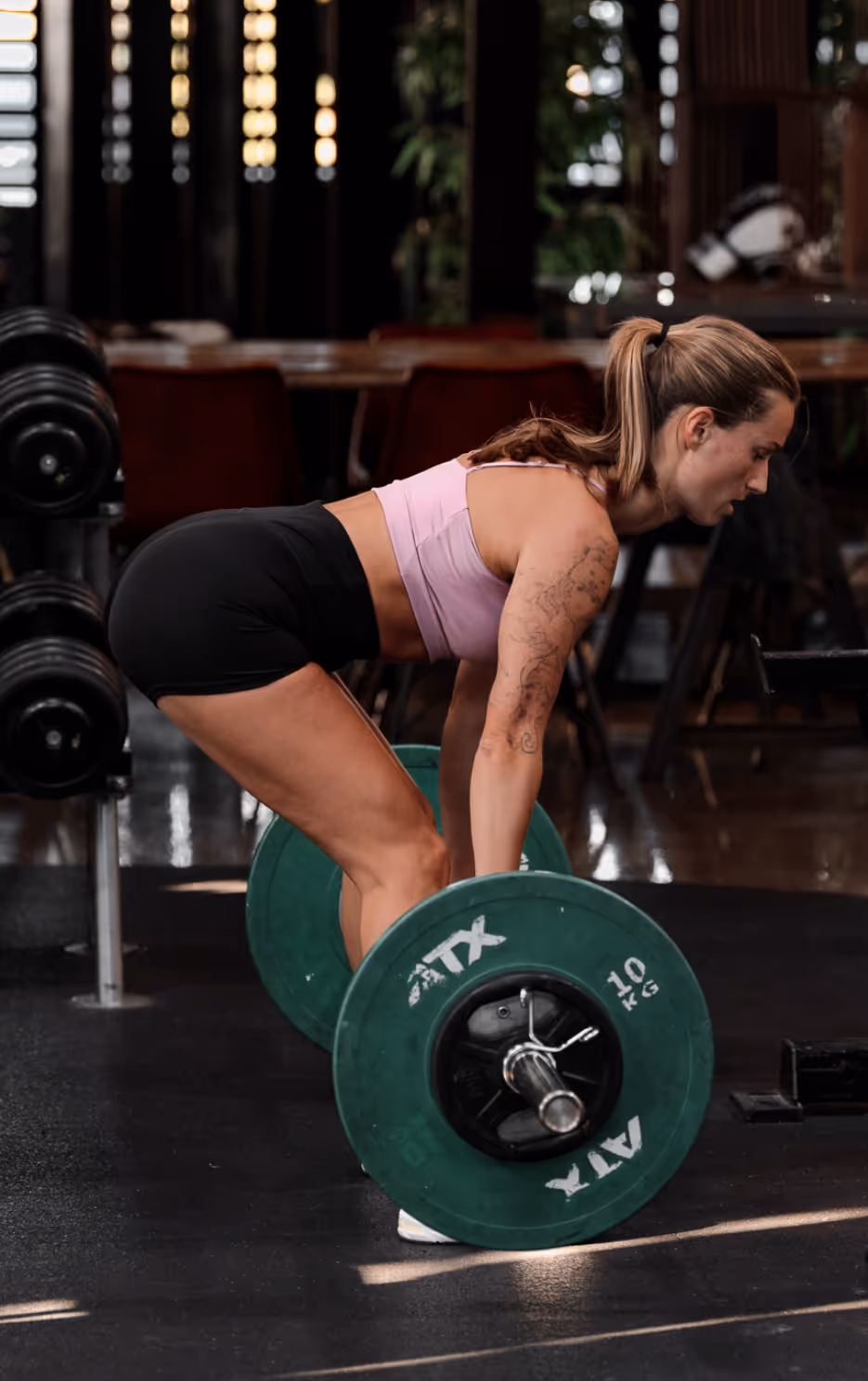 Woman with a ponytail in a gym performing a deadlift with a barbell loaded with 10 kg green weight plates.