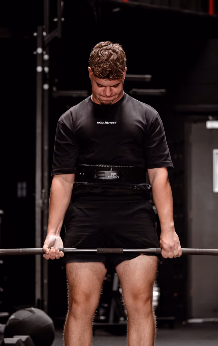 Man wearing a black udp.fitness shirt and lifting a barbell in a gym with a weightlifting belt.
