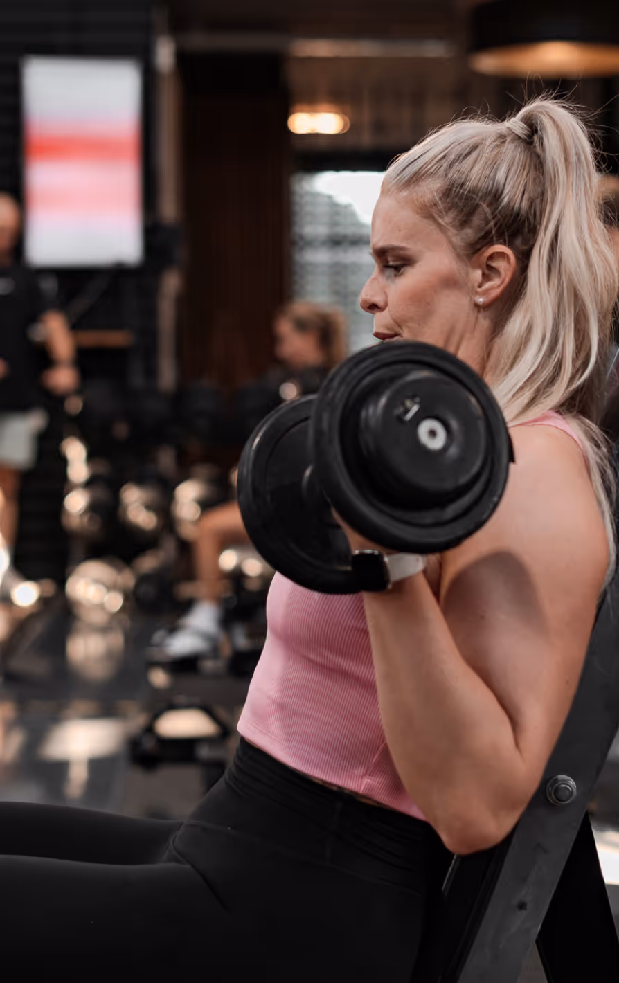 Woman with blonde hair in a ponytail lifting dumbbells while seated on a bench in a gym.