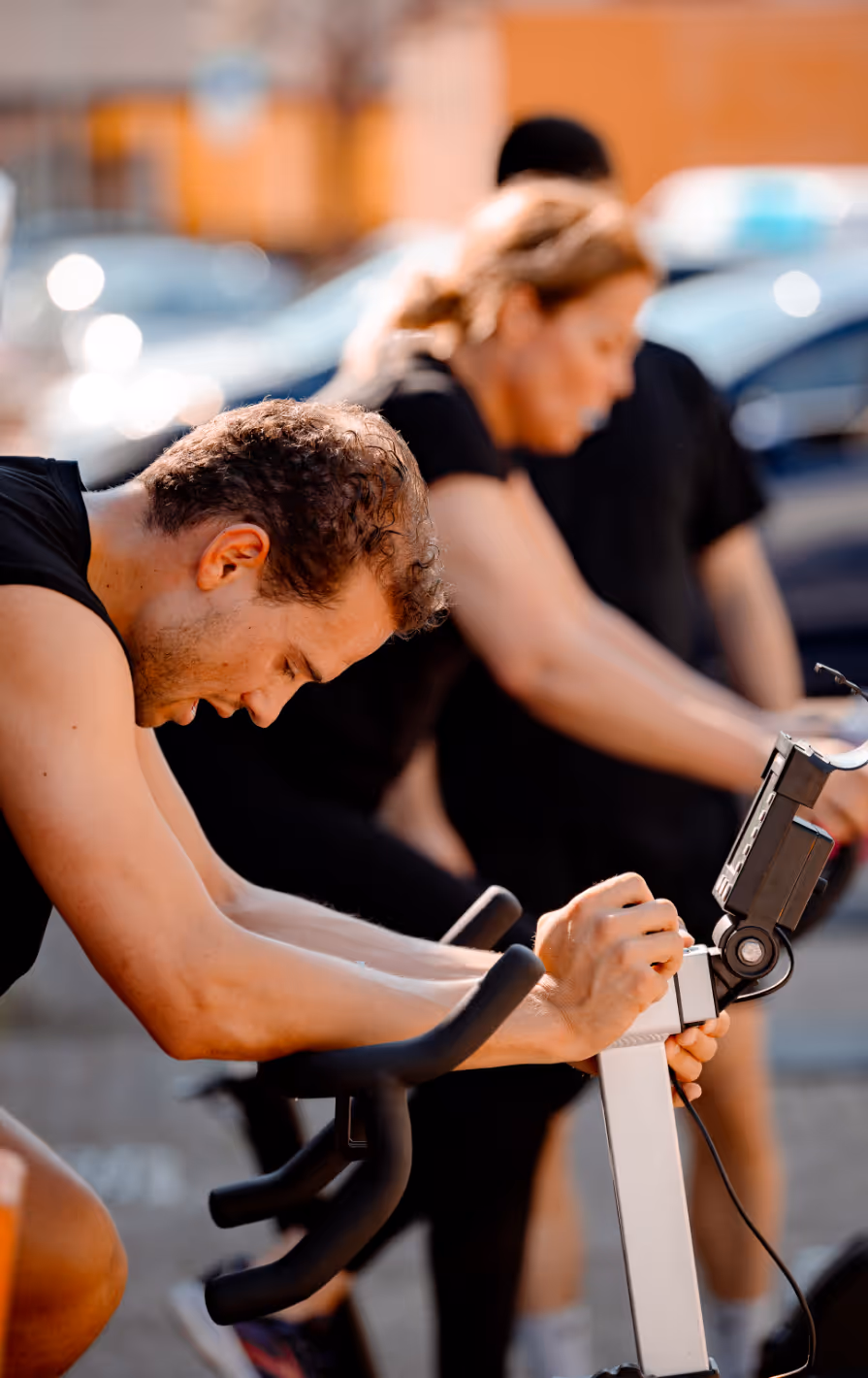 Three people exercising on stationary bikes outdoors, focusing on the man in the foreground.