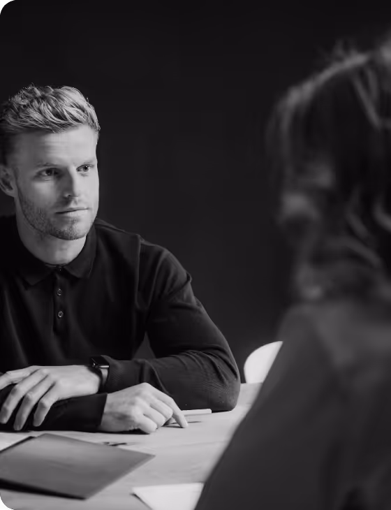 Man with light hair and beard wearing a dark sweater attentively listening to a woman across a table in a black and white photo.