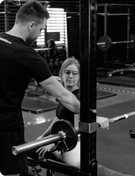 Male trainer assisting a woman with glasses lifting a barbell on a weight bench in a gym.
