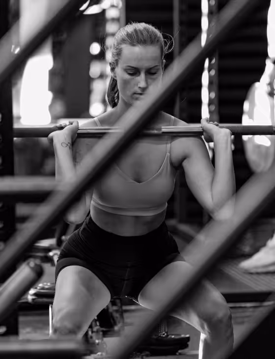 Woman performing a barbell squat in a gym, framed by metal bars in the foreground.