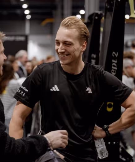 Man with blond hair in black sportswear smiling and holding a water bottle in a crowded indoor gym.