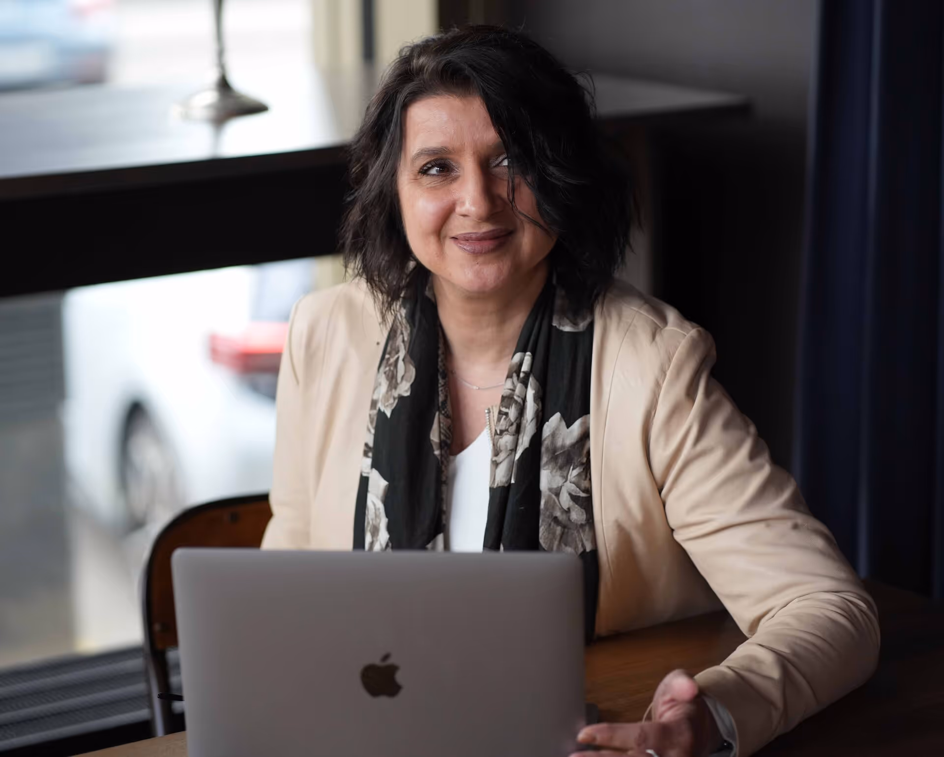 Smiling woman with dark curly hair sitting at a table using a laptop with an Apple logo.
