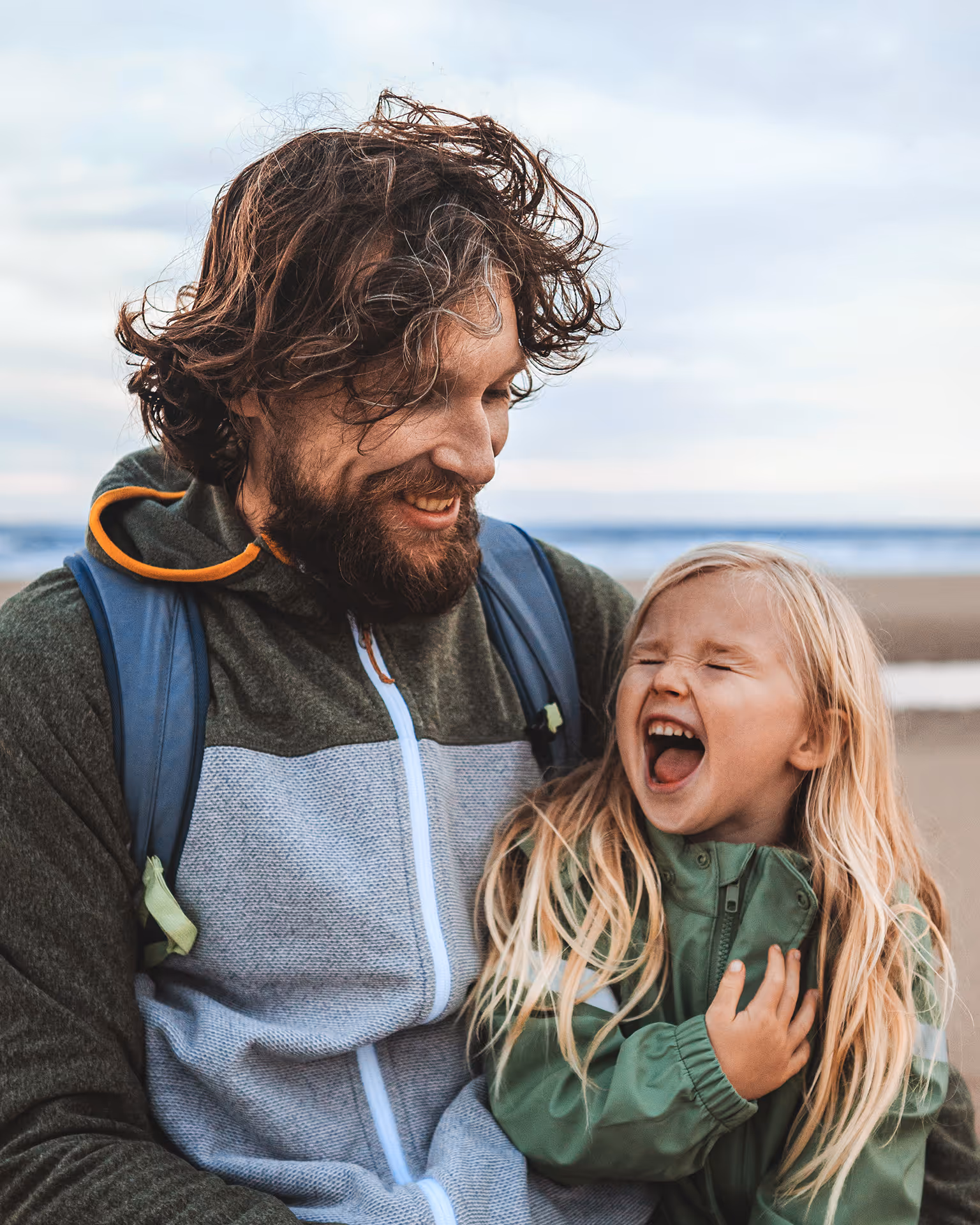 Ein lächelnder Mann mit Bart hält ein lachendes kleines Mädchen mit langen blonden Haaren am Strand.