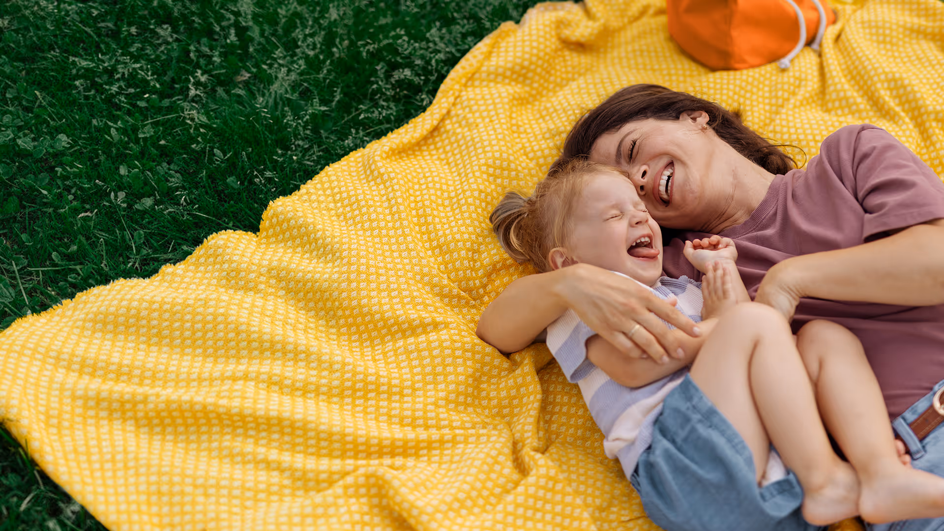 Mutter und Kind lachen gemeinsam auf einer gelben Decke im Freien.