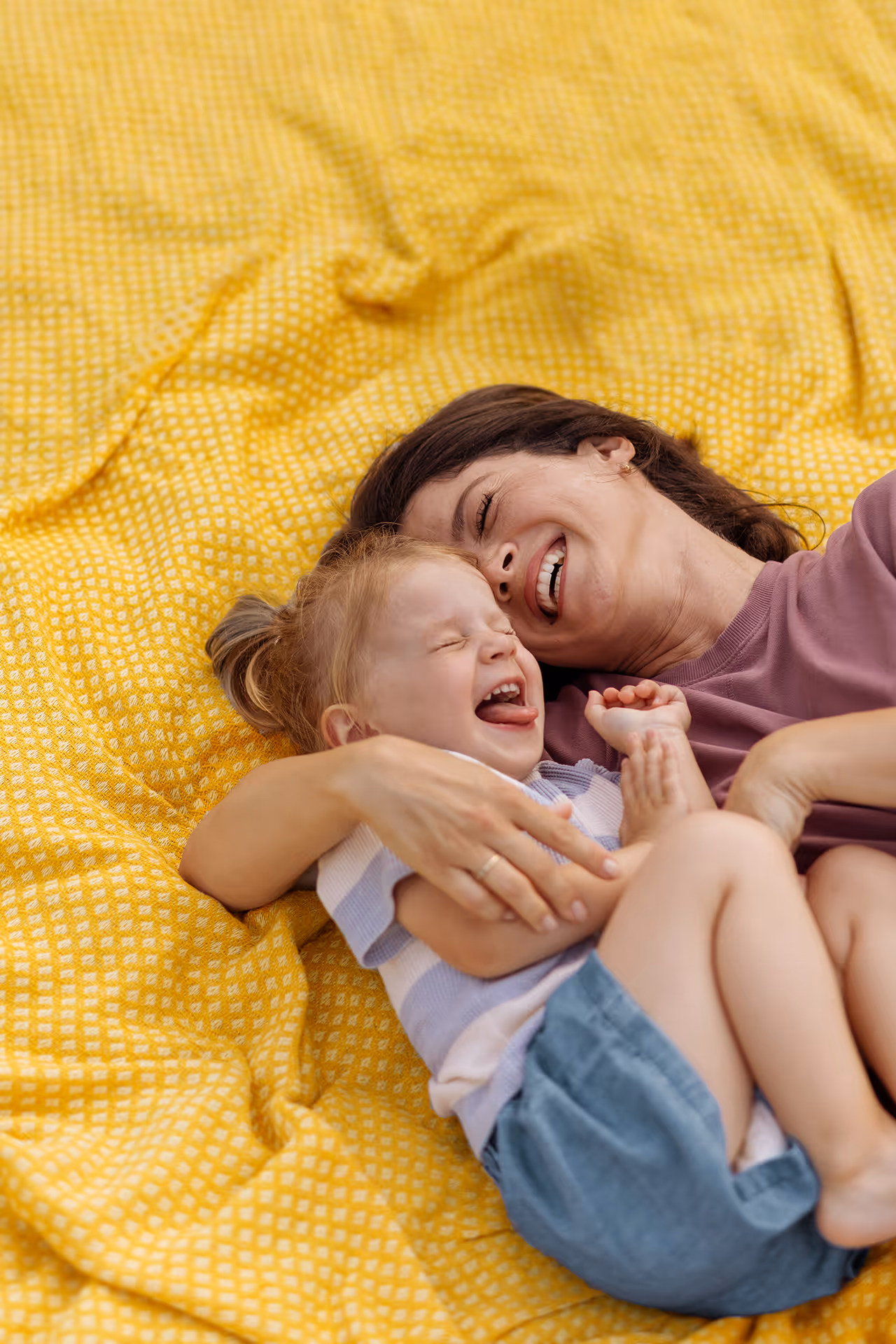 Mutter und Kind lachen gemeinsam auf einer gelben Decke im Freien.