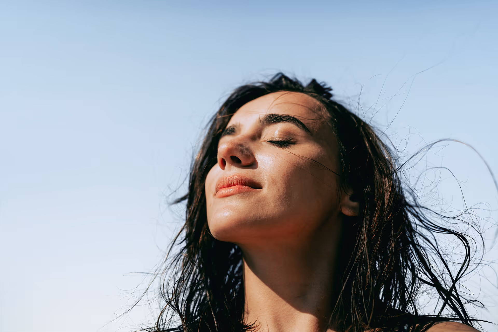 Frau mit geschlossenen Augen und leichtem Lächeln im Sonnenlicht vor klarem Himmel.