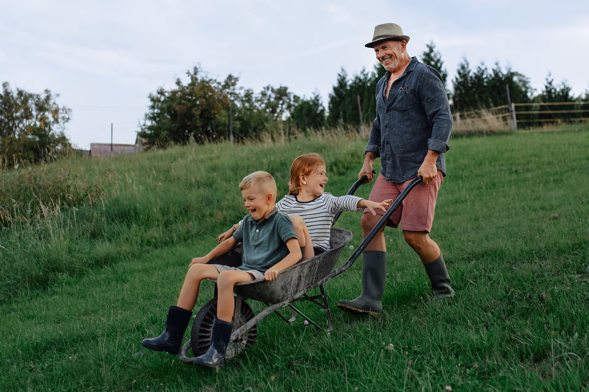 Großvater schiebt zwei lachende Kinder in einer Schubkarre auf einer grünen Wiese.
