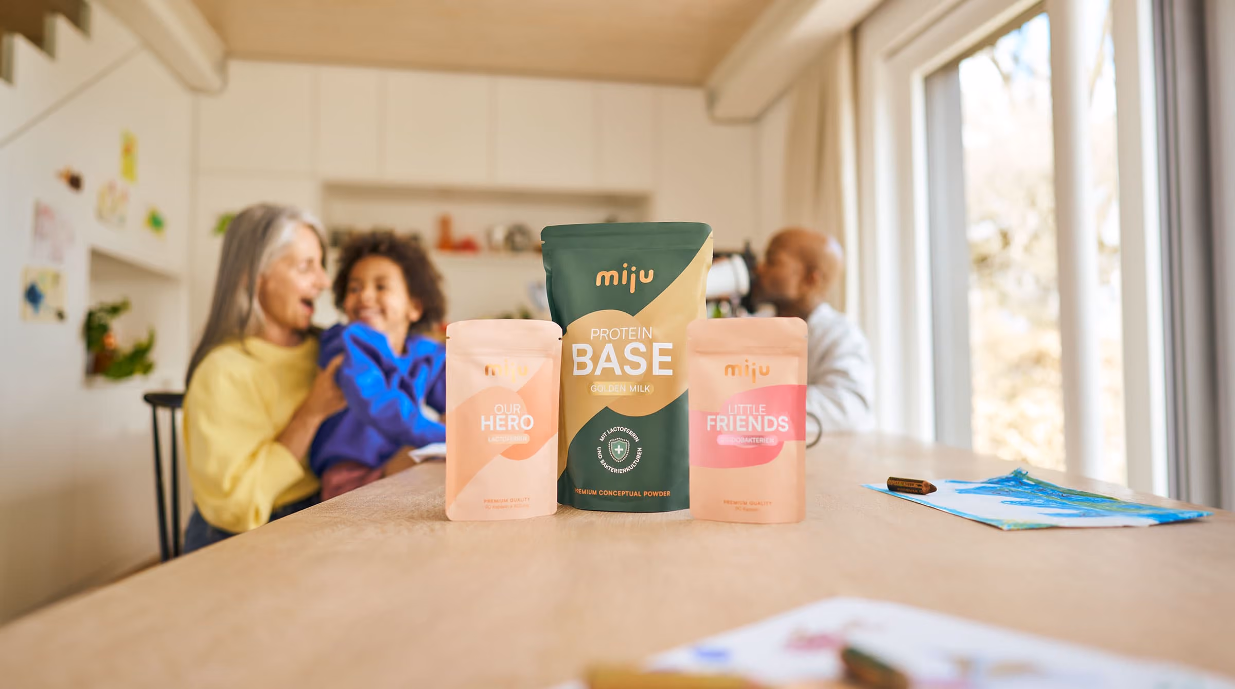 miju Lactoferrin, Bifidobakterien und Proteinpulver vor einer Familie im Alltag. Im Hintergrund trinkt ein Vater einen Proteinshake, während eine Großmutter gemeinsam mit ihrem Enkel lacht.