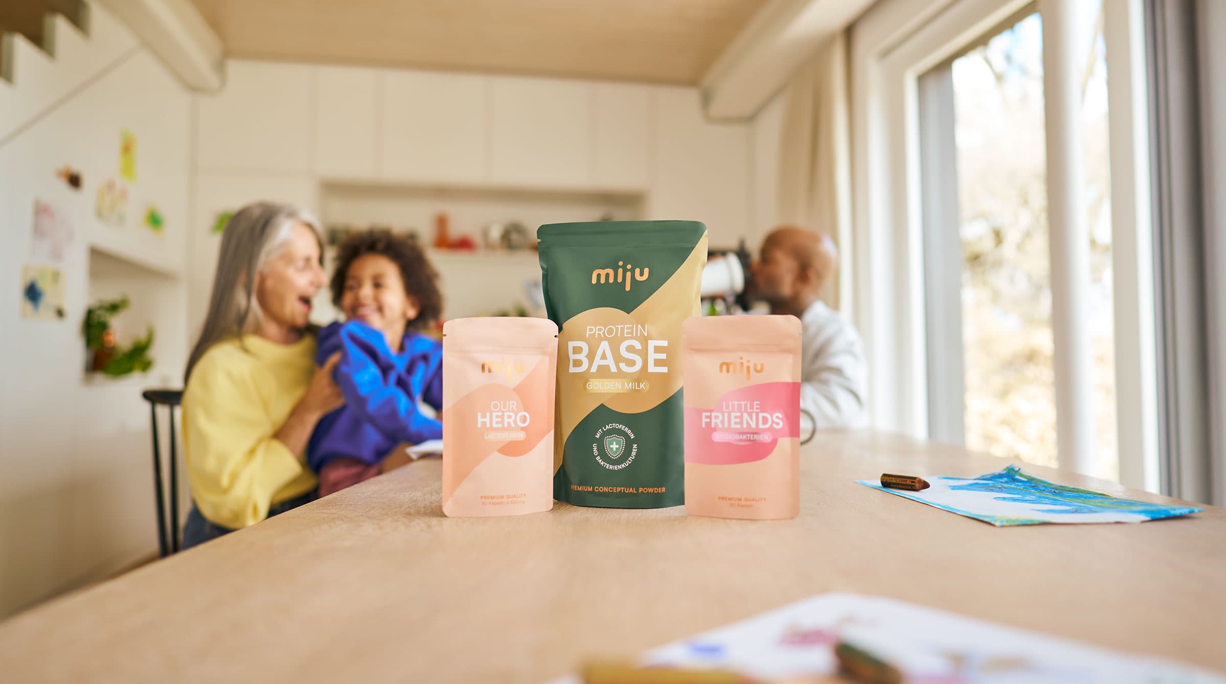 miju Lactoferrin, Bifidobakterien und Proteinpulver vor einer Familie im Alltag. Im Hintergrund trinkt ein Vater einen Proteinshake, während eine Großmutter gemeinsam mit ihrem Enkel lacht.
