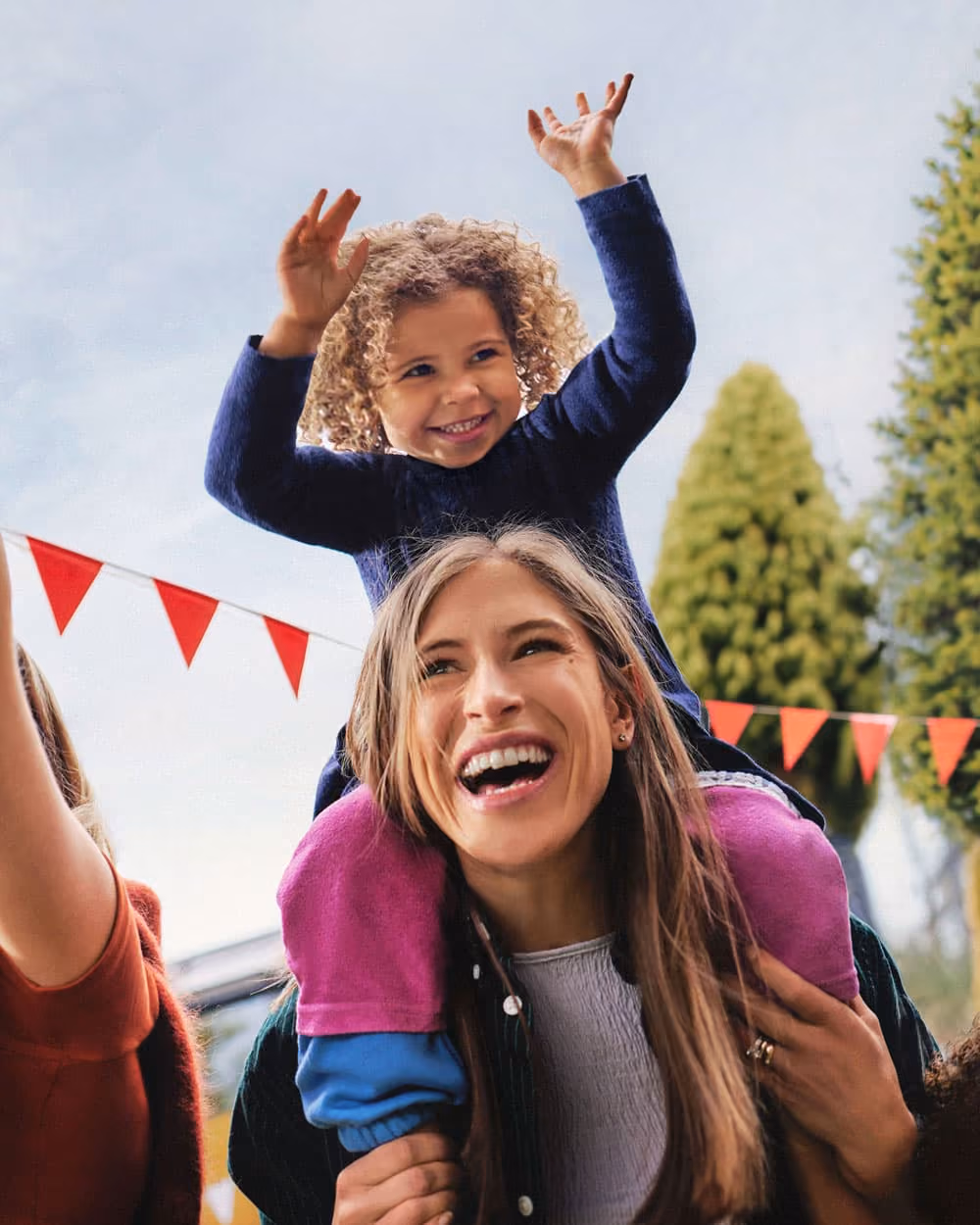 Mutter tanzt auf einer Gartenparty mit ihrer Tochter auf den Schultern. Die fröhliche Szene steht für einen aktiven Lebensstil, Energie und Wohlbefinden im Familienalltag.